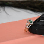 A rose gold ring with a green oval center stone and surrounding clear stones, resting on a peach-colored surface next to a black camera lens.