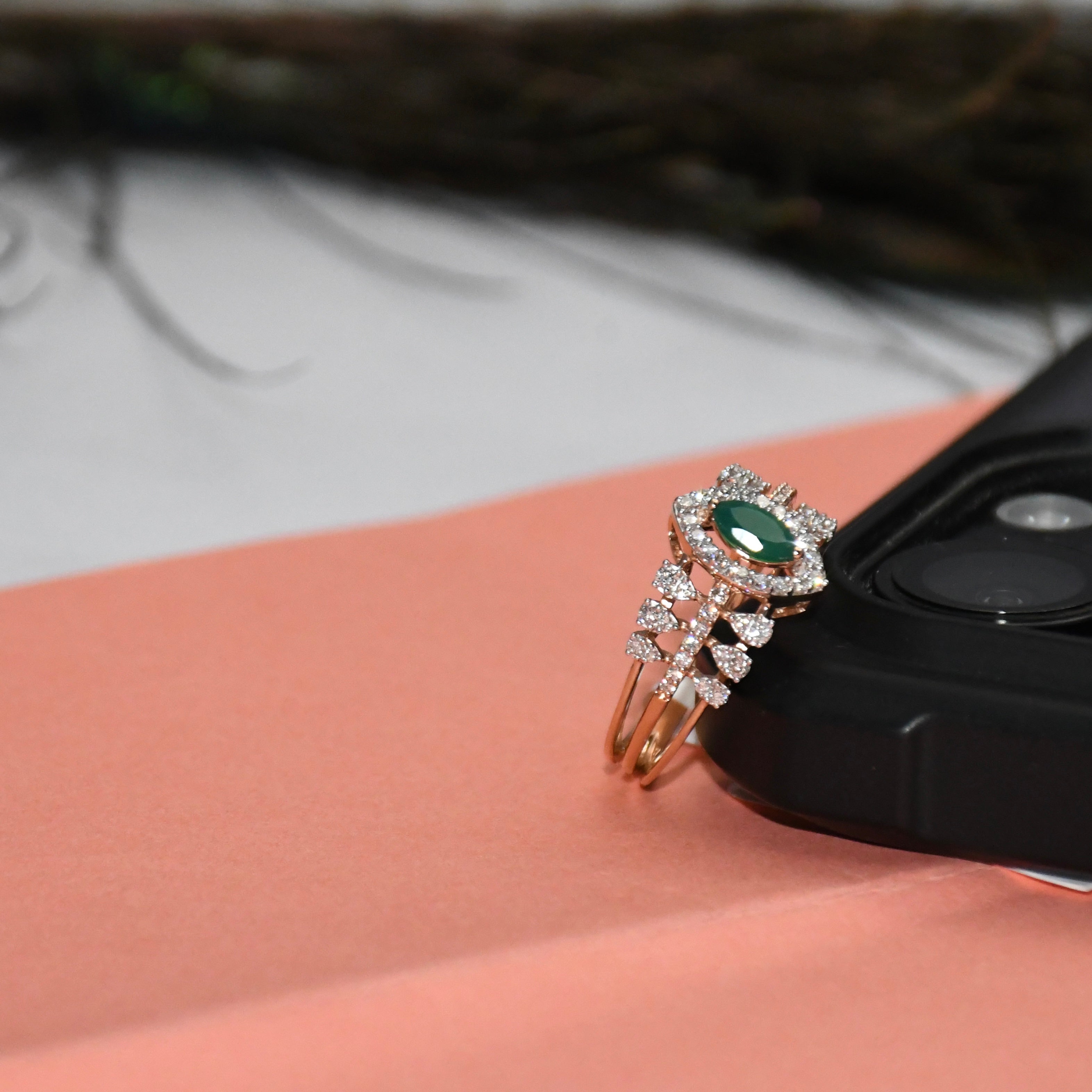 A rose gold ring with a green oval center stone and surrounding clear stones, resting on a peach-colored surface next to a black camera lens.