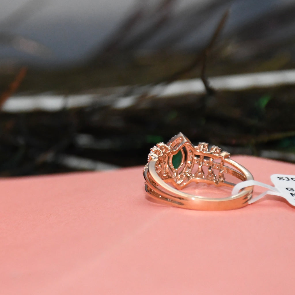 A rose gold ring with a green marquise-cut gemstone and small clear accent stones, resting on a pink surface with a blurred dark background.