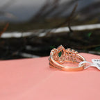 A rose gold ring with a green marquise-cut gemstone and small clear accent stones, resting on a pink surface with a blurred dark background.