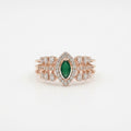 A rose gold ring featuring a central marquise-cut green emerald surrounded by a halo of small clear stones, with additional clear stones along the band, set against a plain white background.