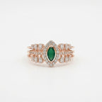 A rose gold ring featuring a central marquise-cut green emerald surrounded by a halo of small clear stones, with additional clear stones along the band, set against a plain white background.