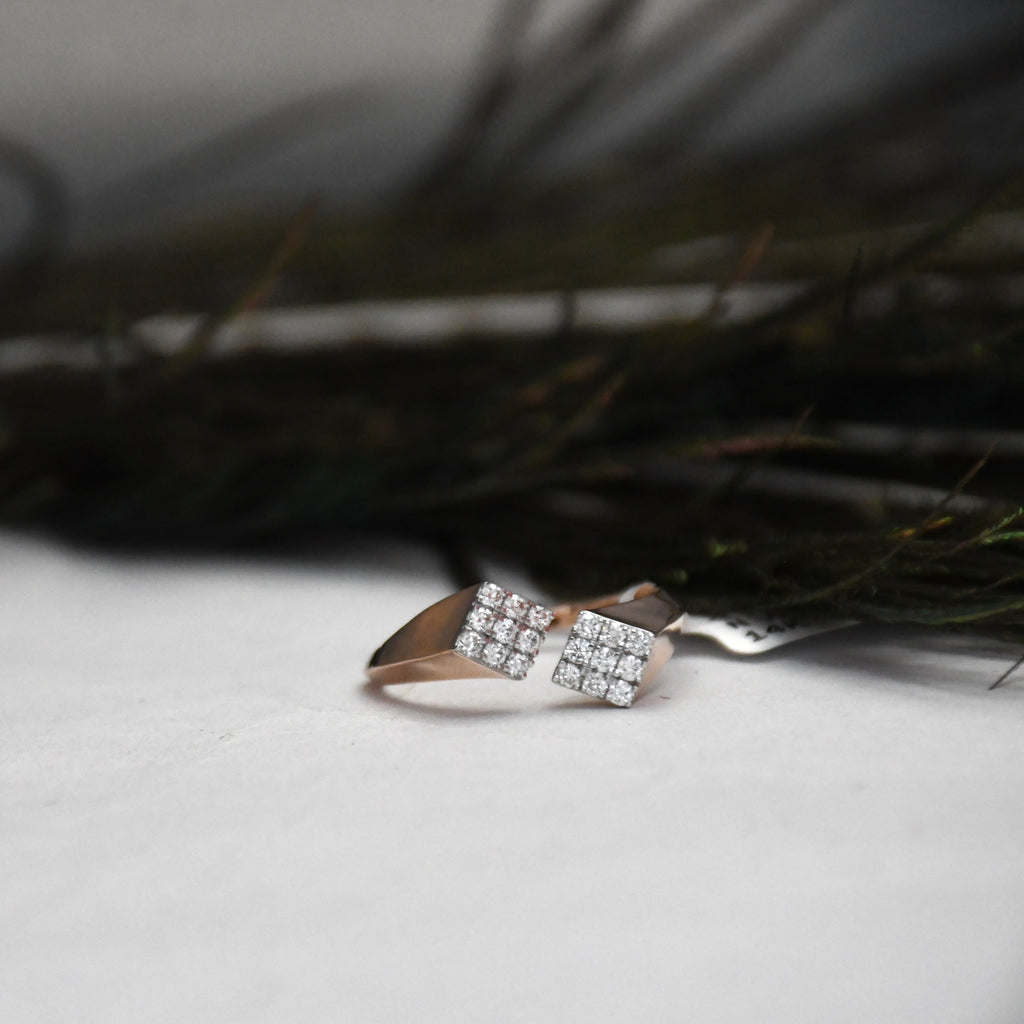 A rose gold ring with two square clusters of clear pavé stones, resting on a white textured surface with dark, blurred foliage in the background.