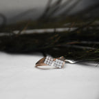 A rose gold ring with two square clusters of clear pavé stones, resting on a white textured surface with dark, blurred foliage in the background.