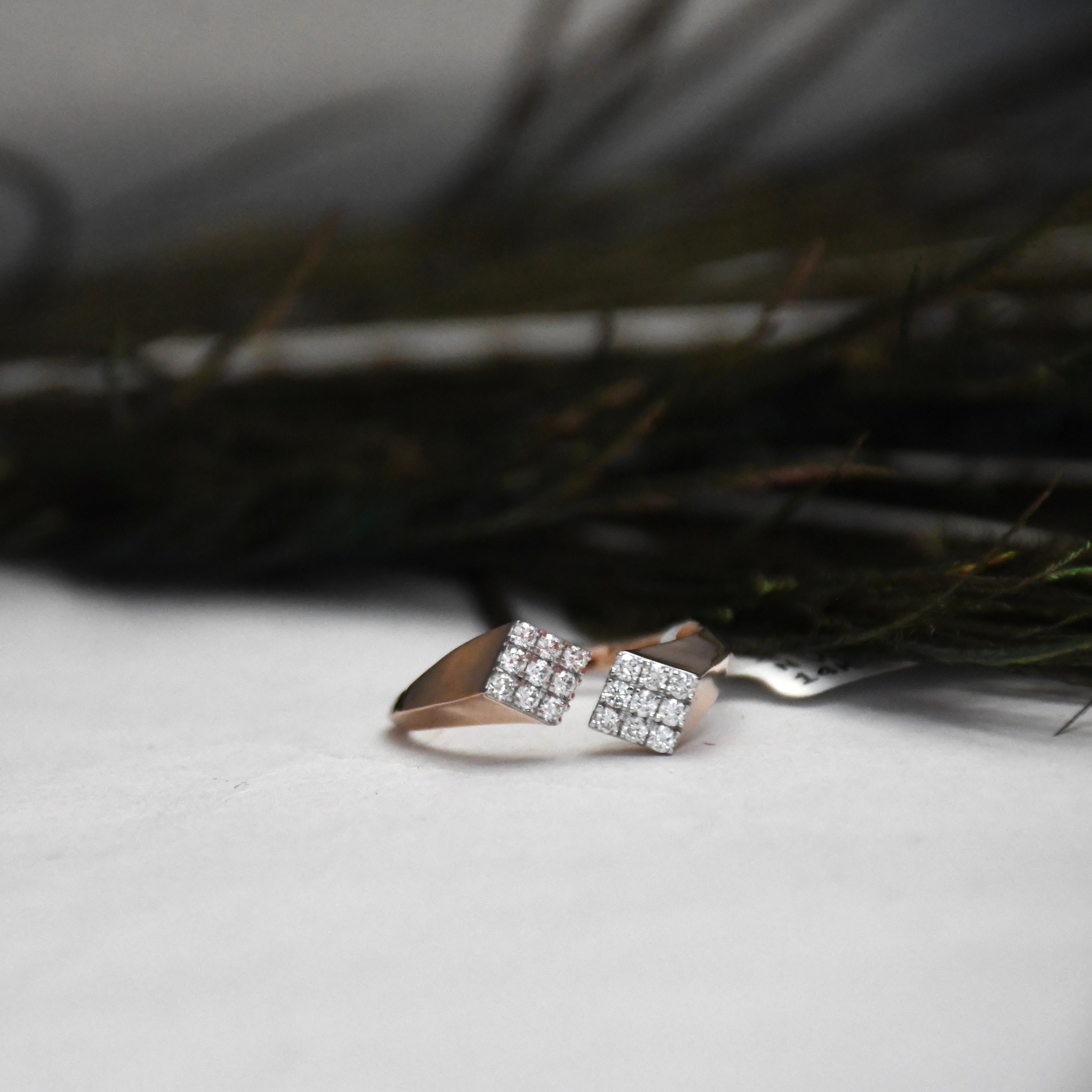 A rose gold ring with two square clusters of clear pavé stones, resting on a white textured surface with dark, blurred foliage in the background.