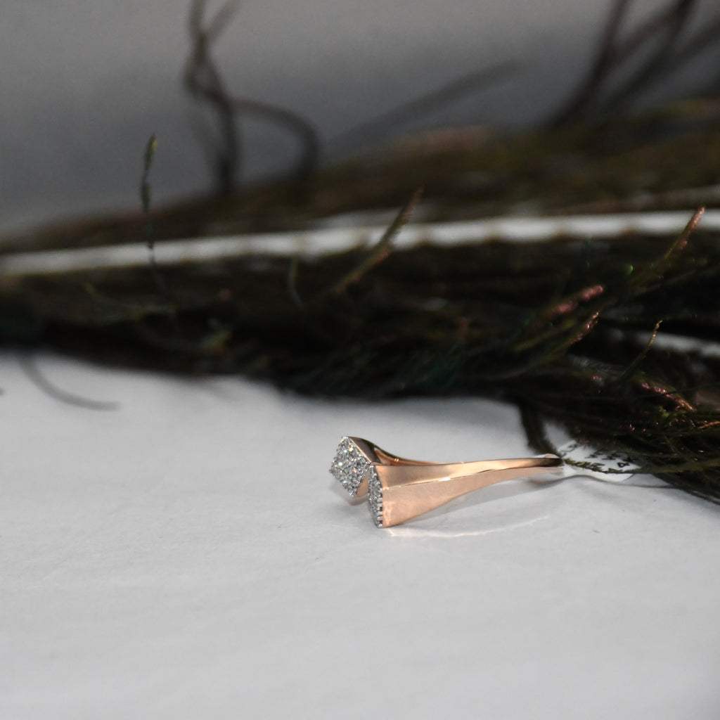 A rose gold ring with clear diamond cluster setting, resting on a white surface with dark, blurred decorative elements in the background.