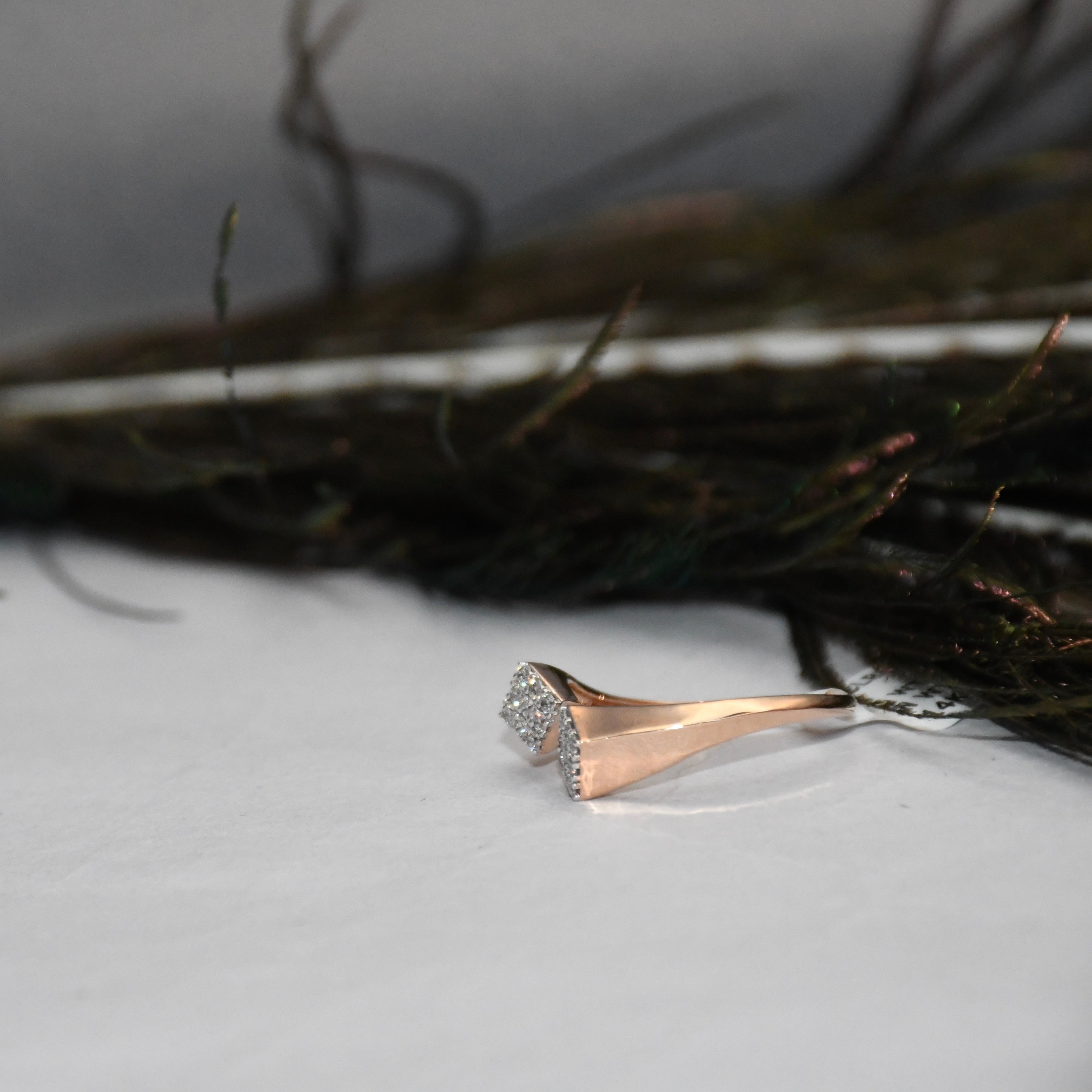 A rose gold ring with clear diamond cluster setting, resting on a white surface with dark, blurred decorative elements in the background.