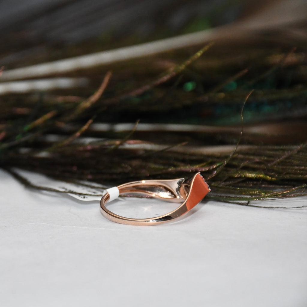A rose gold ring with a small, abstract curved stone set in a white band, resting on a white surface with dark feathers in the background.