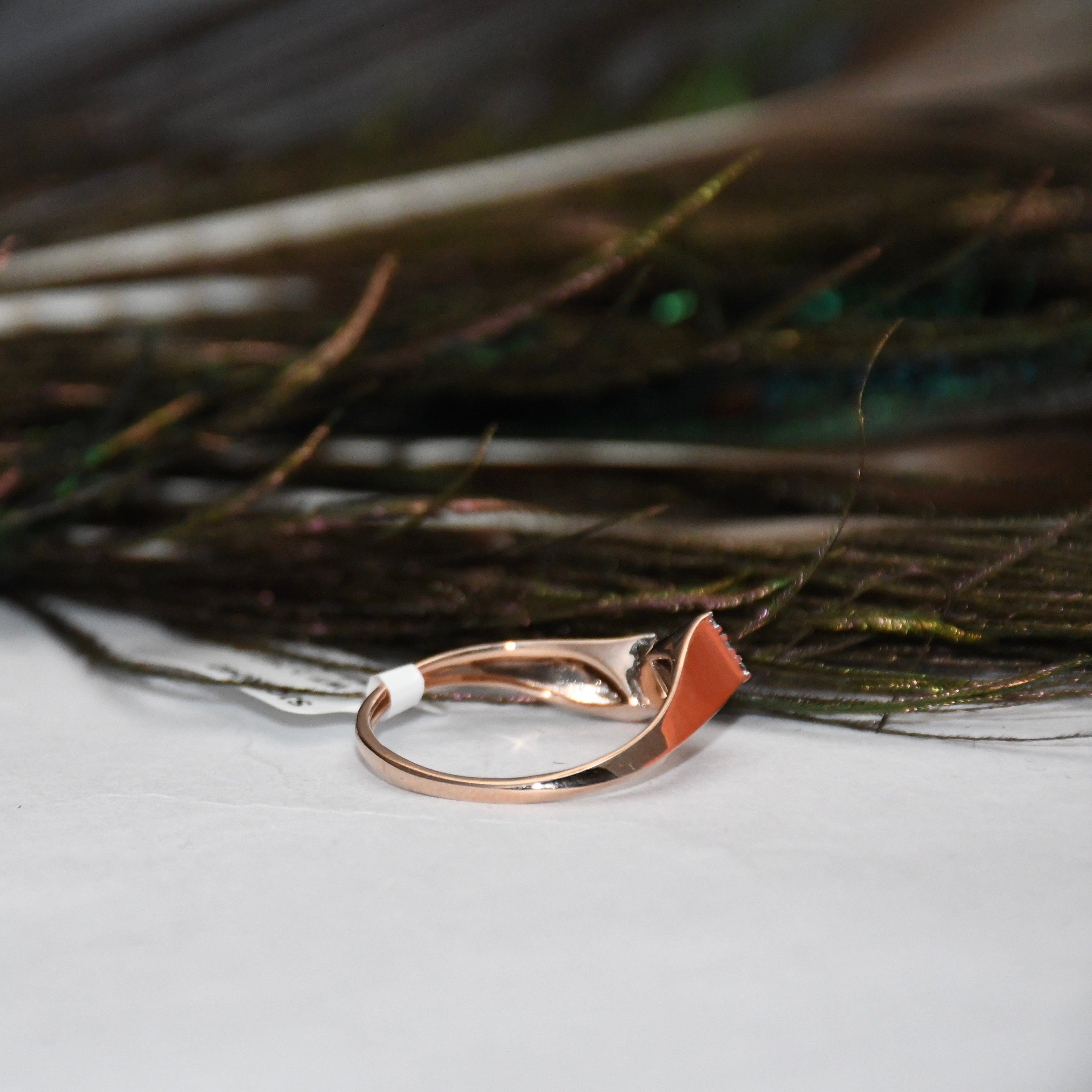A rose gold ring with a small, abstract curved stone set in a white band, resting on a white surface with dark feathers in the background.