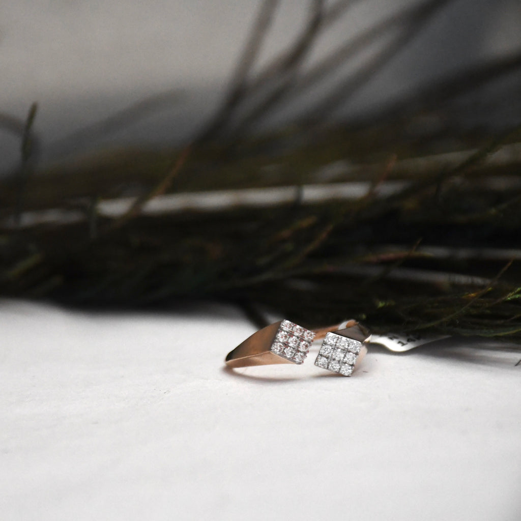 A close-up of a ring with two square pavé-set clear stones on a rose gold band, resting on a white surface with dark blurred branches in the background.