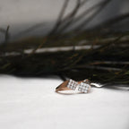A close-up of a ring with two square pavé-set clear stones on a rose gold band, resting on a white surface with dark blurred branches in the background.