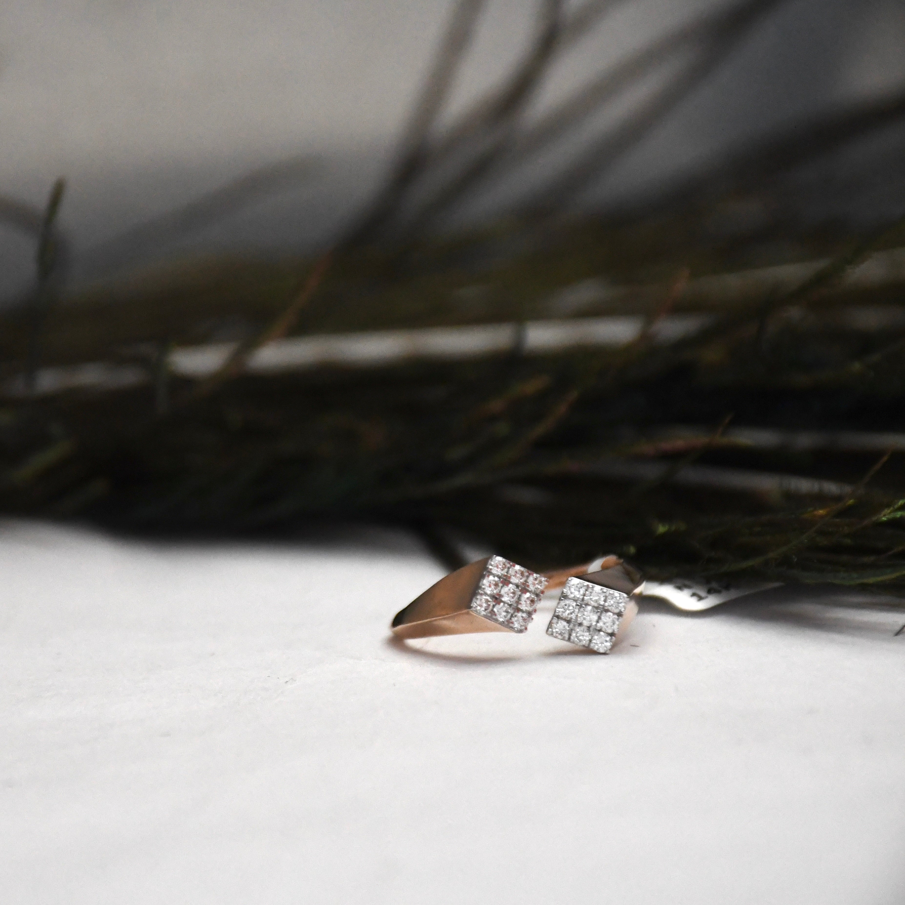 A close-up of a ring with two square pavé-set clear stones on a rose gold band, resting on a white surface with dark blurred branches in the background.