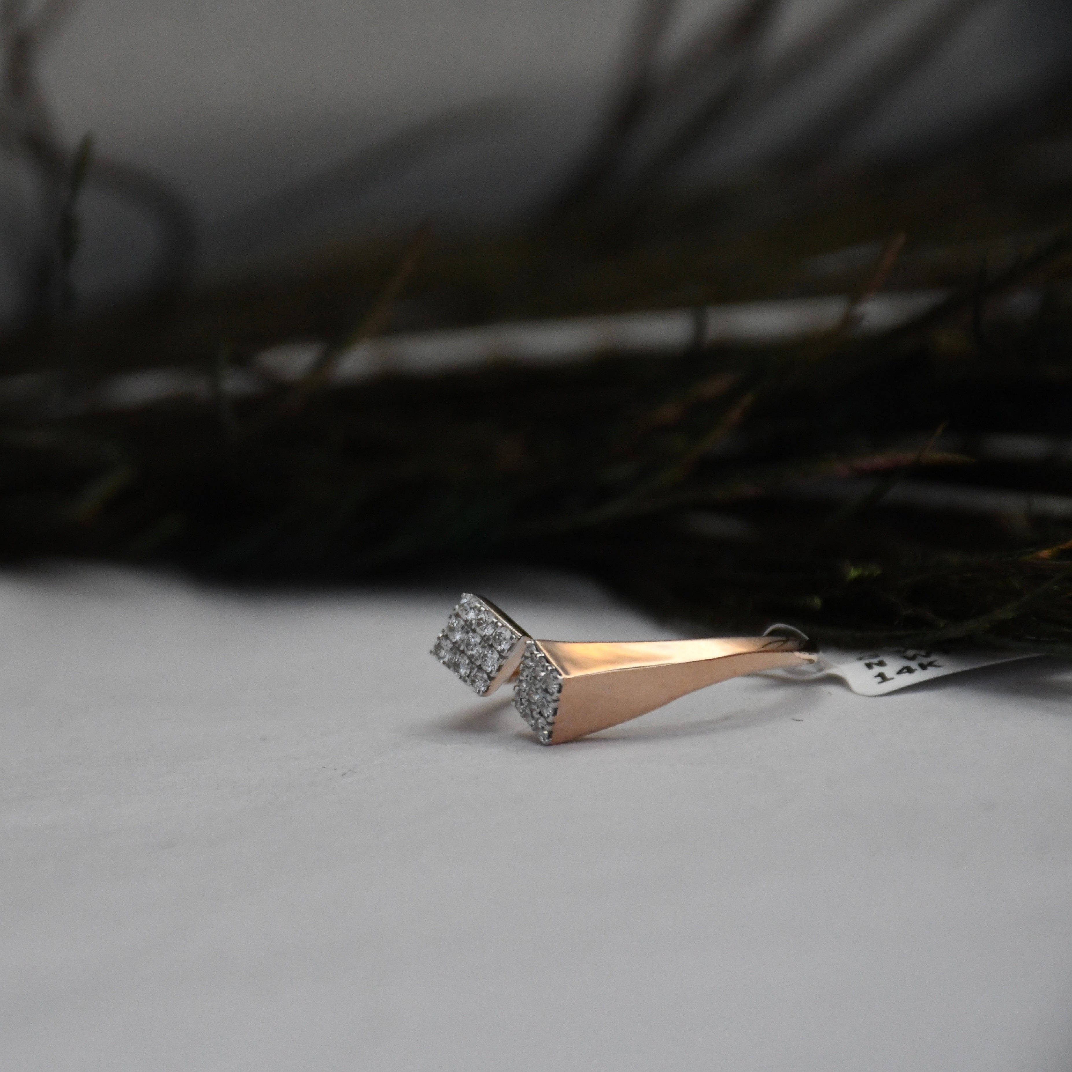 A modern geometric ring featuring a tapered rose gold band with a pavé-set square-shaped diamond cluster on the front, resting on a light gray surface with a dark, blurred background.