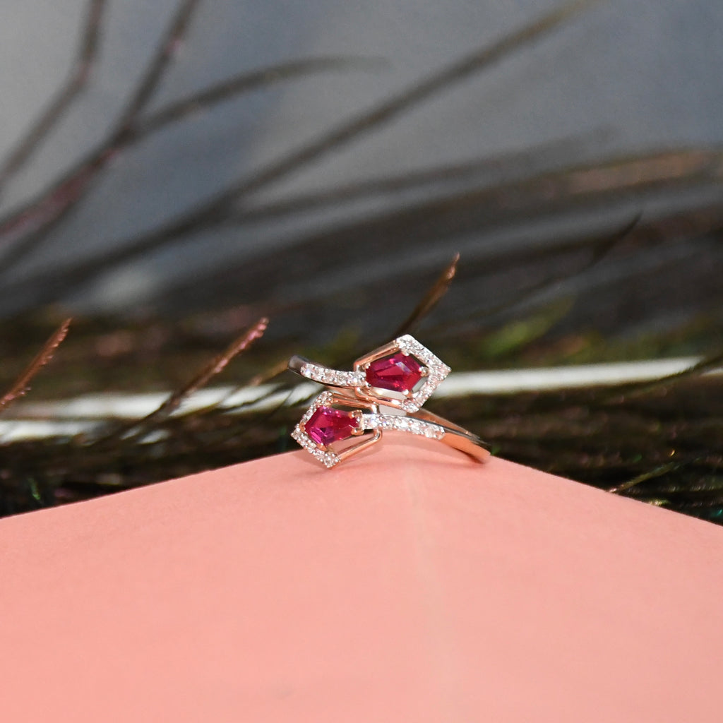 A rose gold ring with two pink gemstones and small clear accent stones, resting on a pink surface with a blurred dark background.