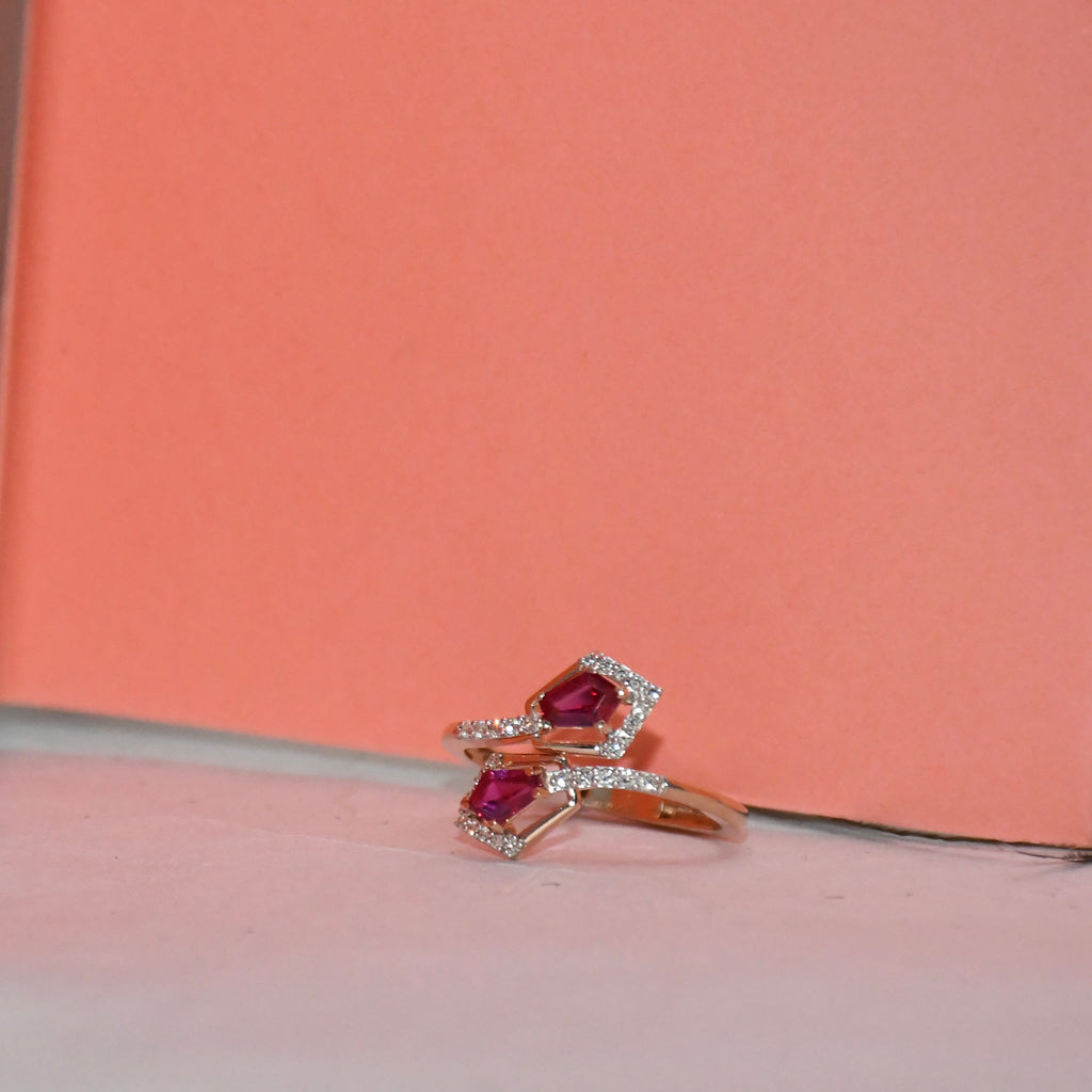 A gold ring with two pinkish-red gemstones surrounded by small clear stones, resting on a light surface against a peach background.