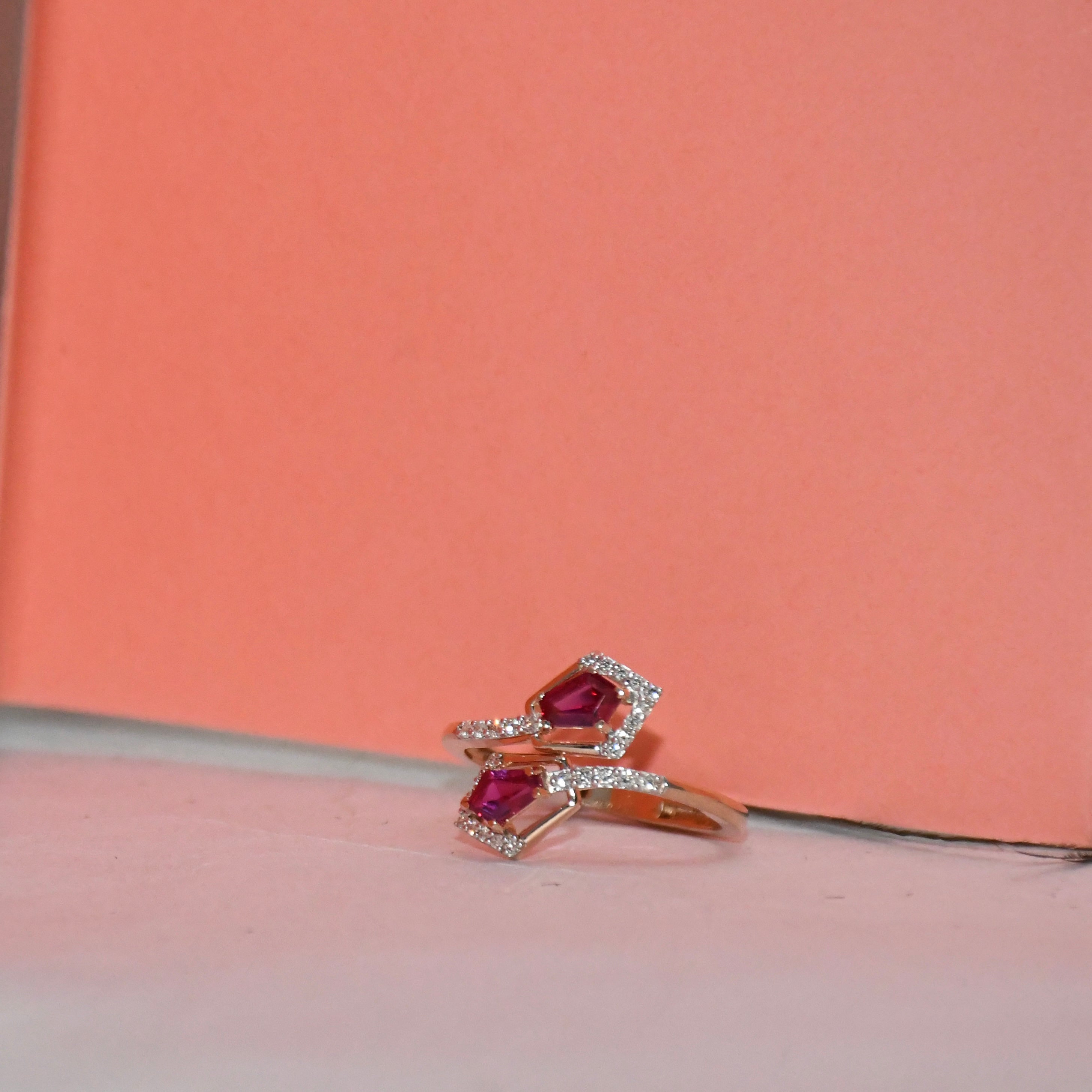 A gold ring with two pinkish-red gemstones surrounded by small clear stones, resting on a light surface against a peach background.