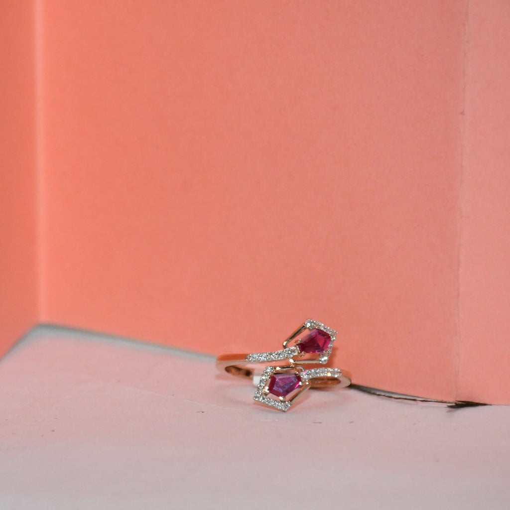 A close-up of a rose gold ring with two pink gemstones and small clear accent stones, resting on a light surface against a peach-pink background.