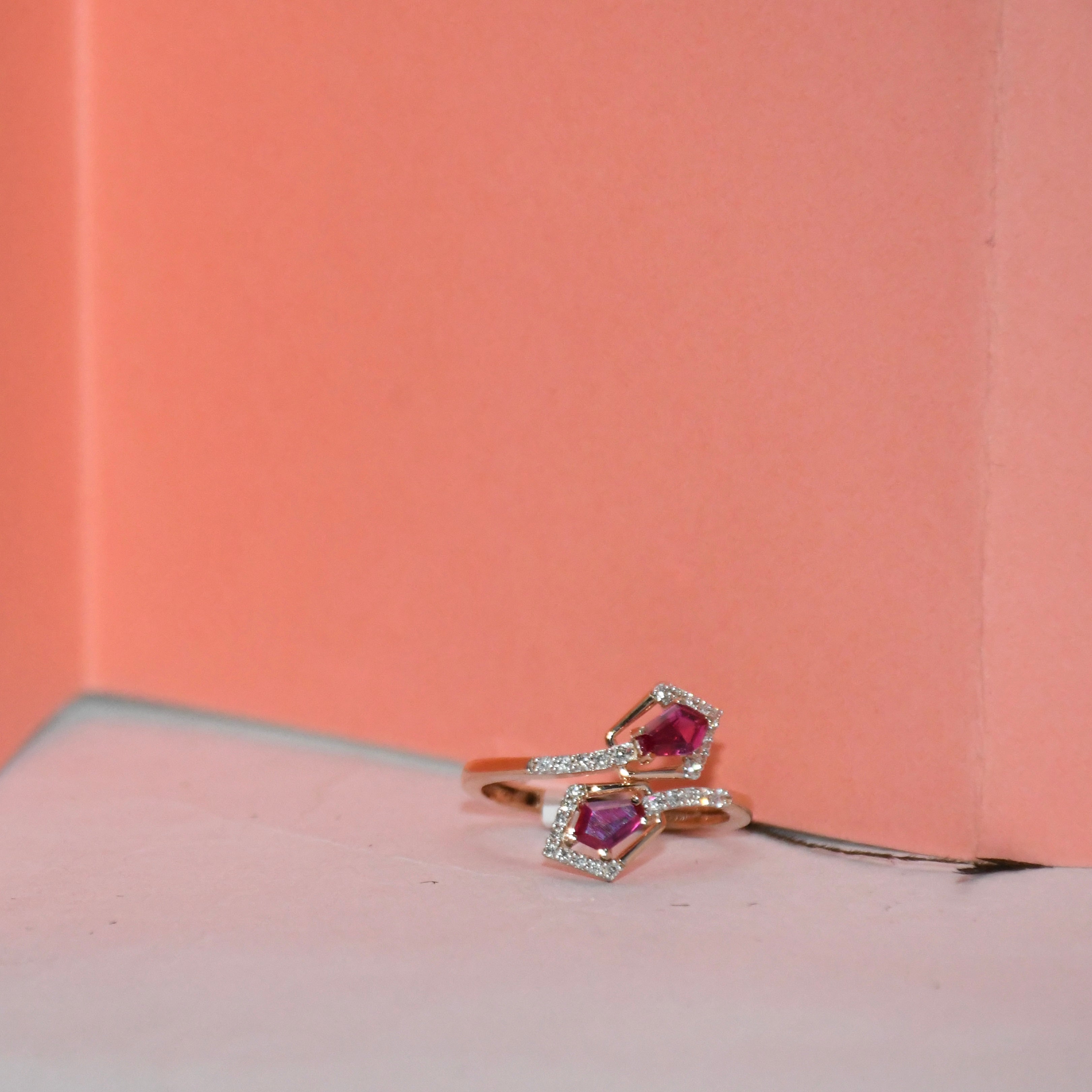 A close-up of a rose gold ring with two pink gemstones and small clear accent stones, resting on a light surface against a peach-pink background.