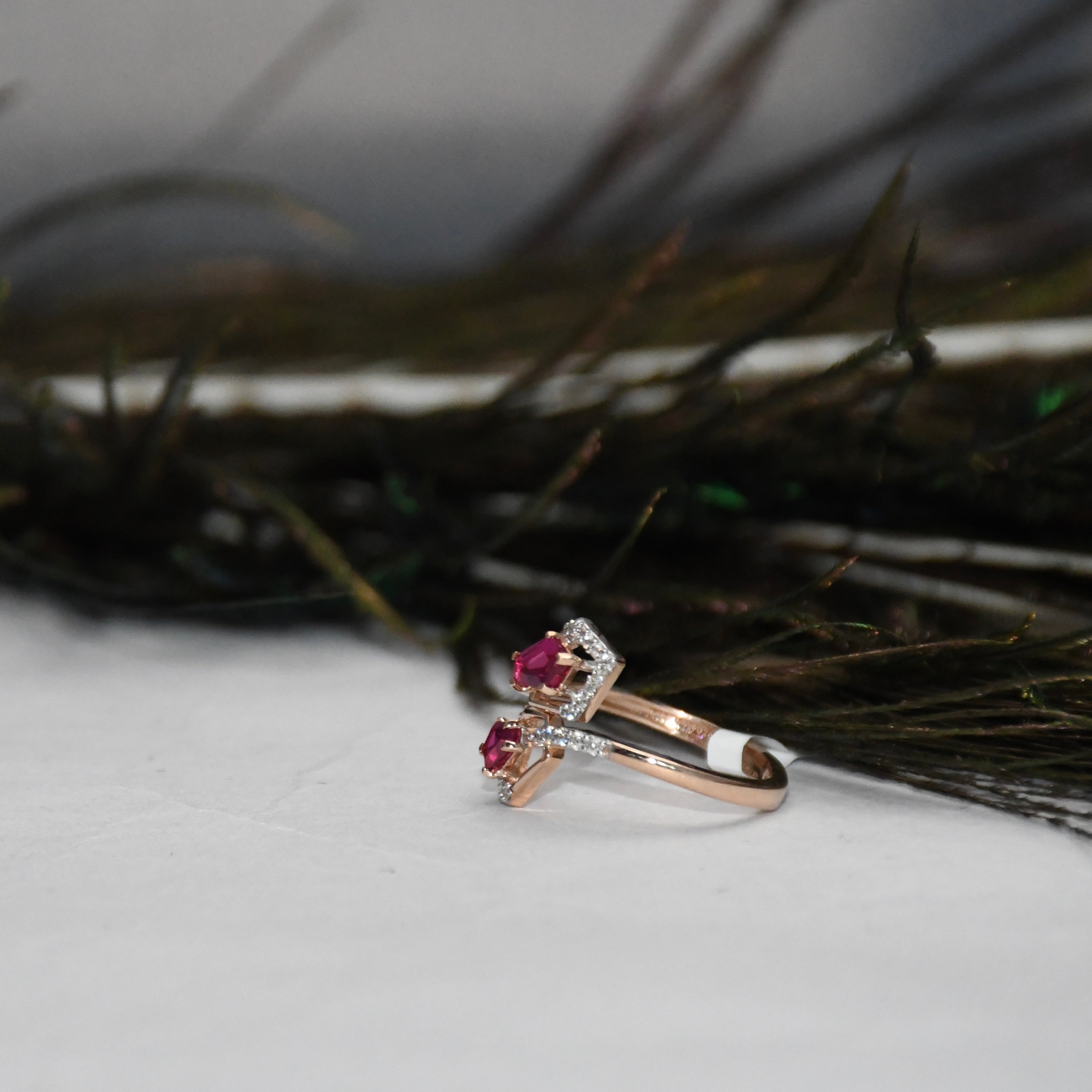 A rose gold ring with two pink gemstones and small clear accent stones, displayed on a white surface with dark decorative feathers in the background.