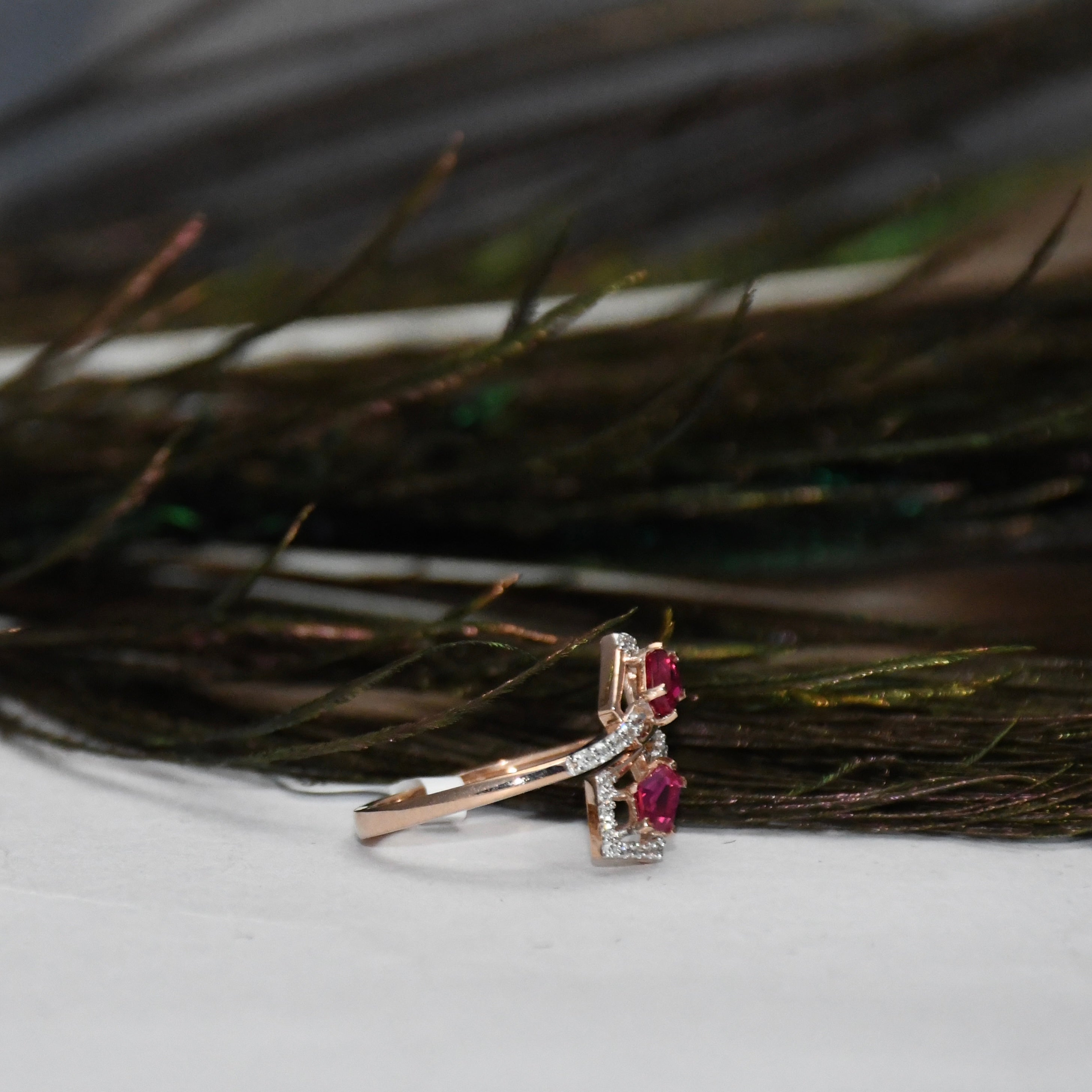 A rose gold ring with two pinkish-red gemstones and small clear accent stones, displayed on a white surface with dark feathers in the background.