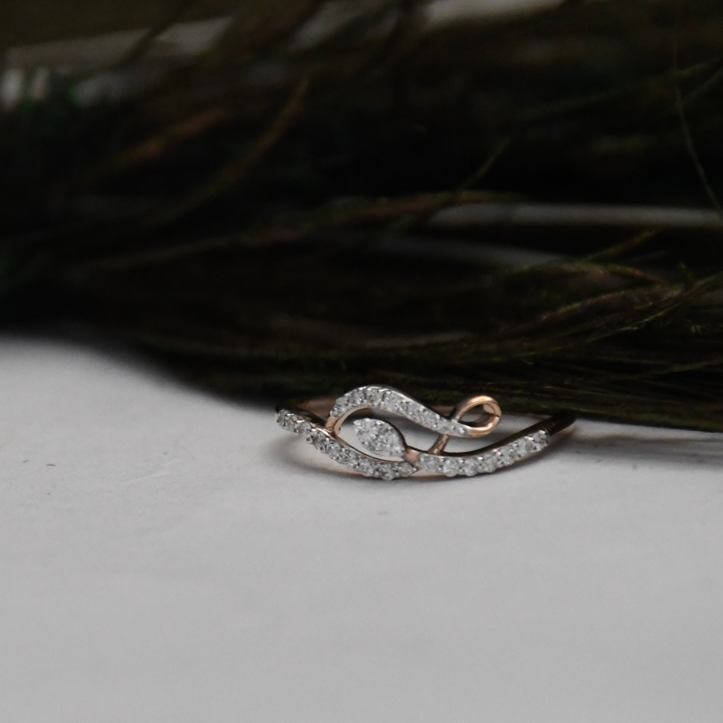 A delicate ring with a curved, leaf-like design paved with clear stones, featuring rose gold accents on a white surface with dark feathers in the background.