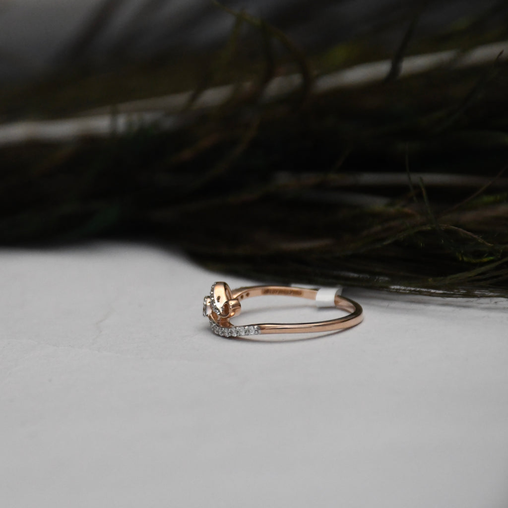 A delicate rose gold ring with a small floral cluster and a pavé diamond band, resting on a light gray surface with dark feathers in the background.