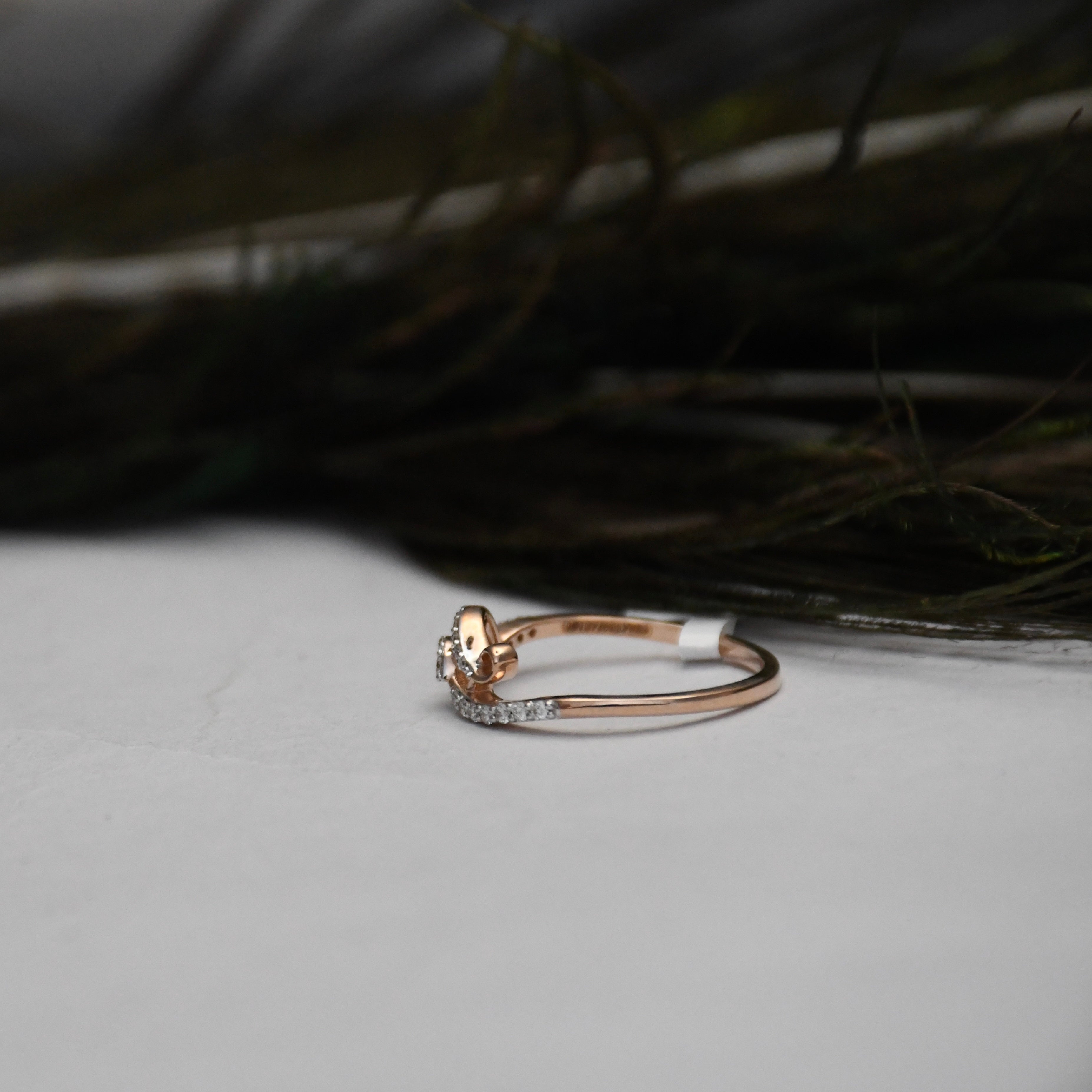 A delicate rose gold ring with a small floral cluster and a pavé diamond band, resting on a light gray surface with dark feathers in the background.