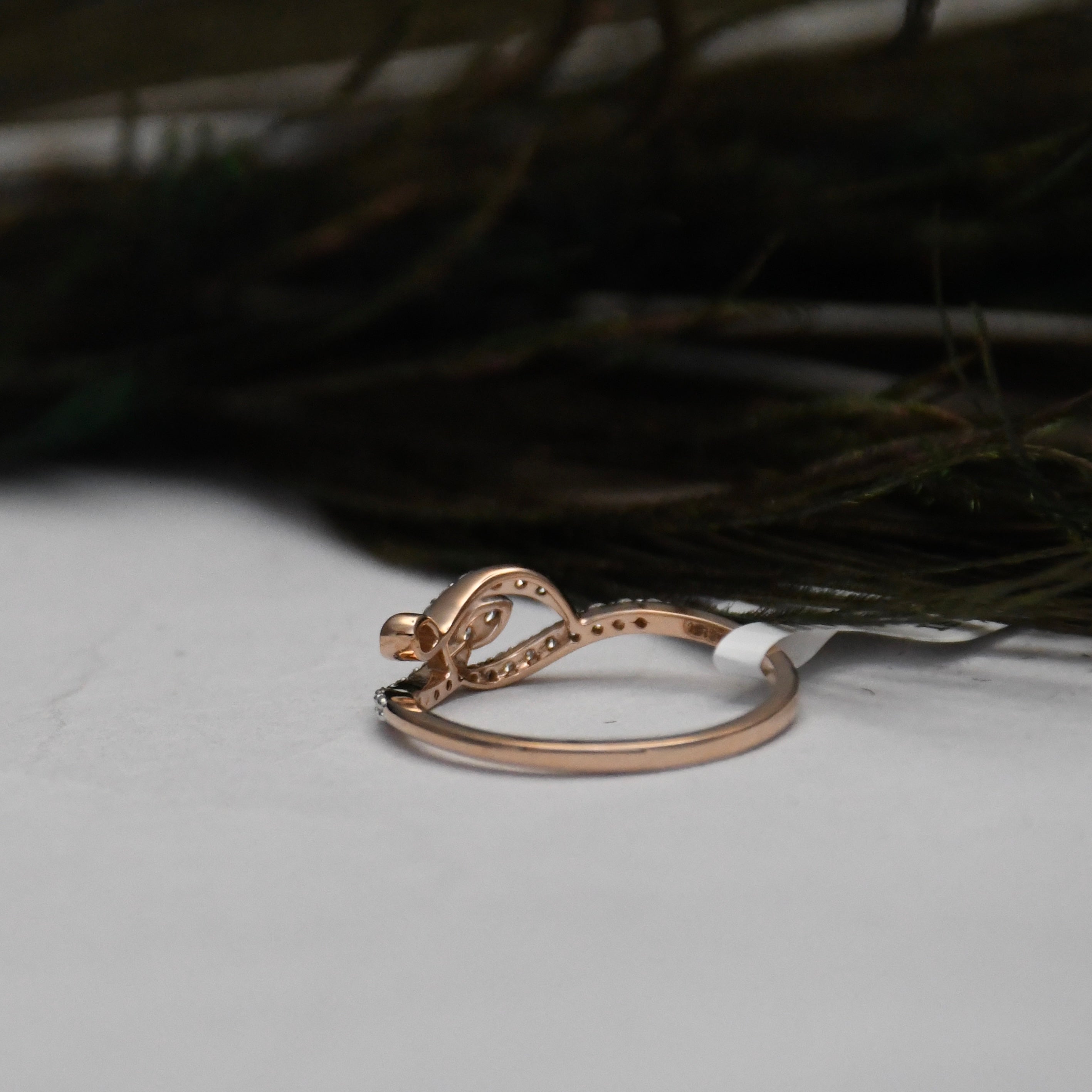 A rose gold ring with a decorative bow-like top and small pavé-set stones, shown on a white surface with dark feathers in the background.
