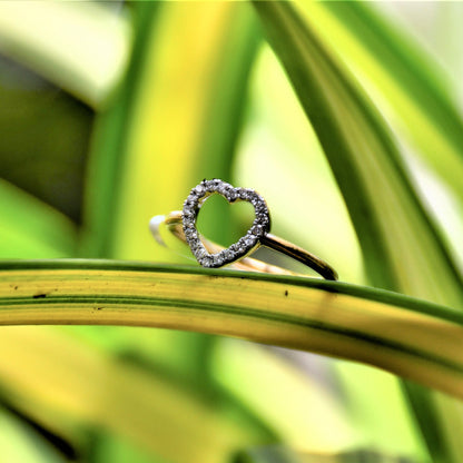 Delicate heart-shaped diamond ring set on a thin gold band, placed on green and yellow variegated leaves in natural light.