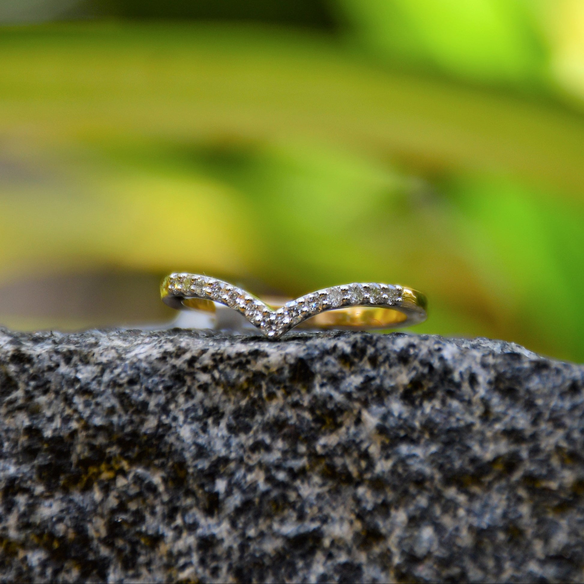 A gold ring featuring a V-shaped diamonds centerpiece, set on a rocky surface with a blurred green background.