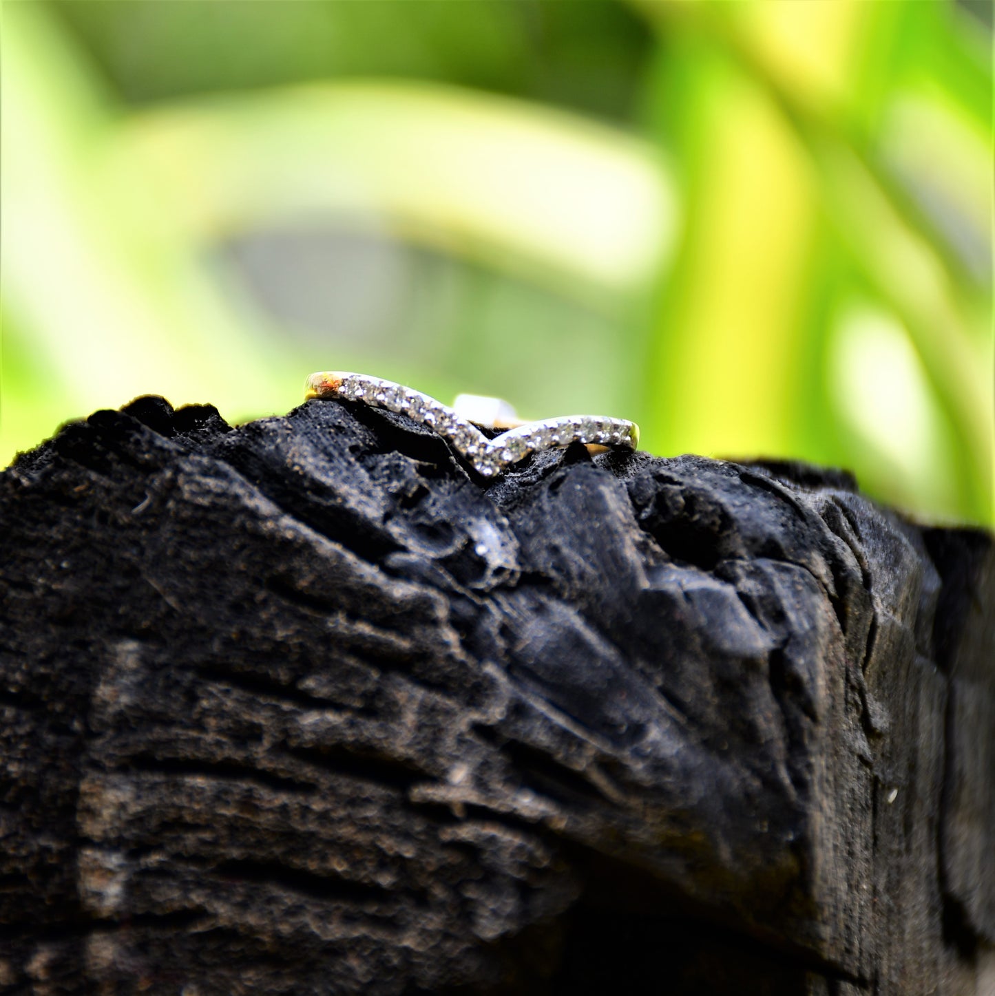 A gold ring featuring a V-shaped diamonds centerpiece, set on a rocky surface with a blurred green background.