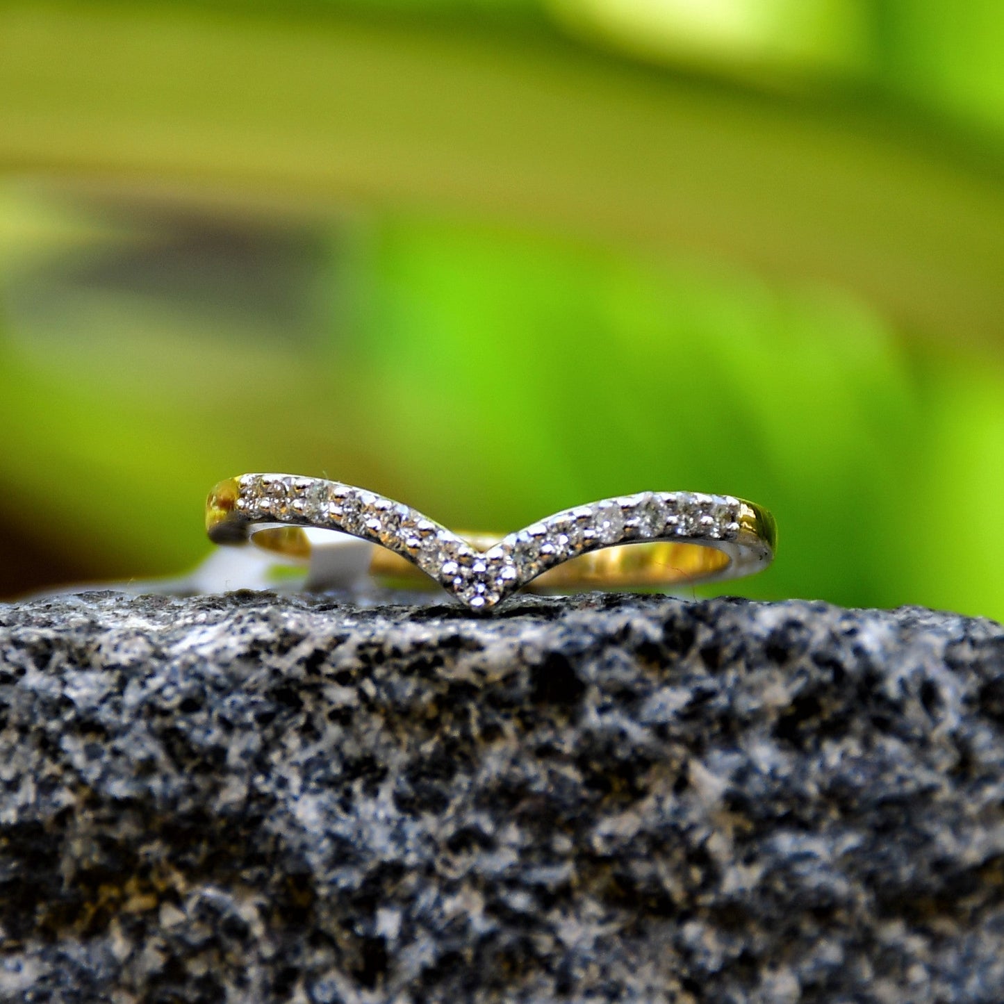 A gold ring featuring a V-shaped diamonds centerpiece, set on a rocky surface with a blurred green background.