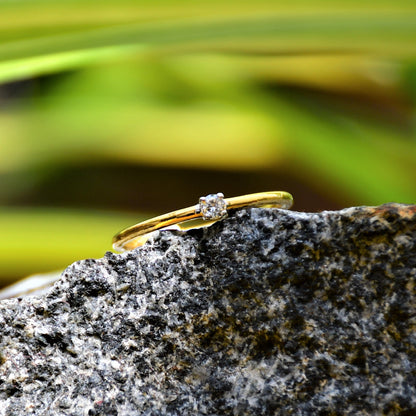A close-up image of a delicate gold ring with a single small diamond solitaire set at the center, displayed on a textured black stone surface with soft green leaves in the blurred background, creating a natural and elegant composition.