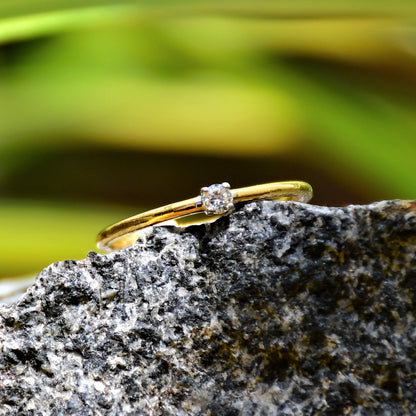 A close-up image of a delicate gold ring with a single small diamond solitaire set at the center, displayed on a textured black stone surface with soft green leaves in the blurred background, creating a natural and elegant composition.