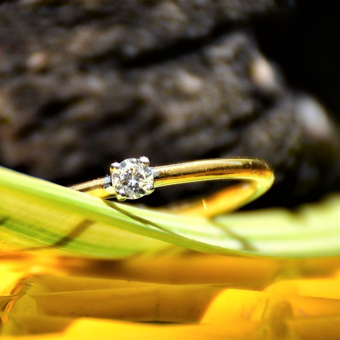 A close-up image of a delicate gold ring with a single small diamond solitaire set at the center, displayed on a yellow surface with soft green leaves in the blurred background, creating a natural and elegant composition.