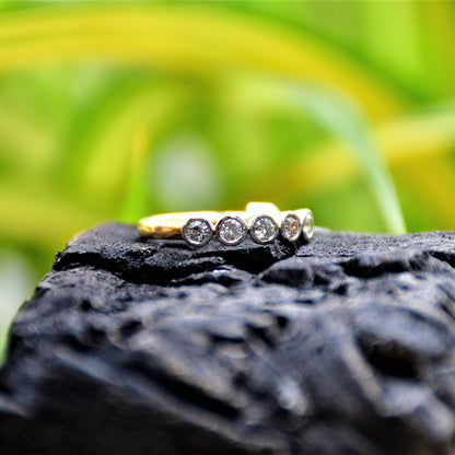 A yellow gold band ring with a series of set diamonds, featuring smaller diamonds, all set in a solid gold band, displayed against a light yellow and green background and on a black rough surface.