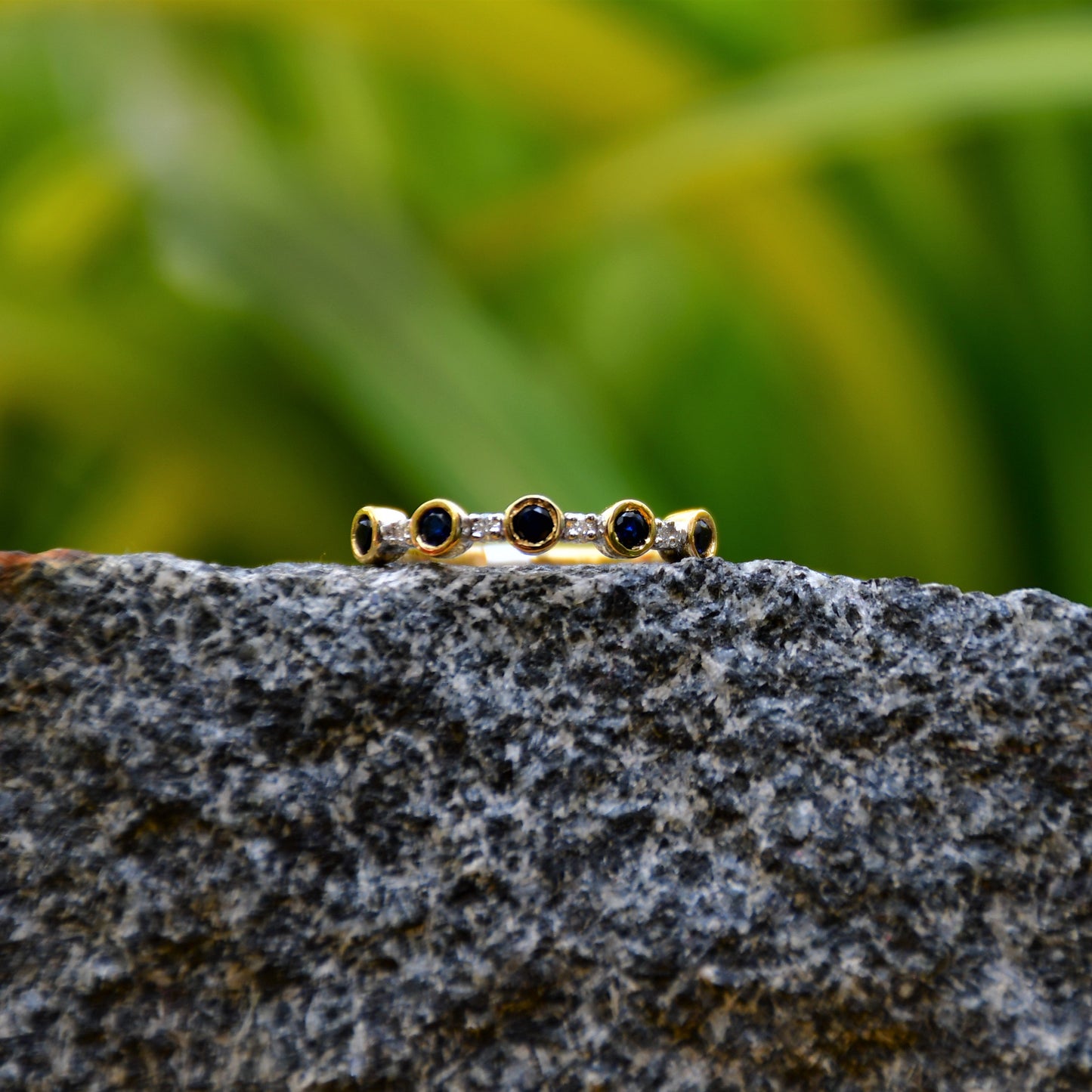 A 14kt gold diamond band ring with blue color stones, positioned on a rocky surface with green foliage in the background.
