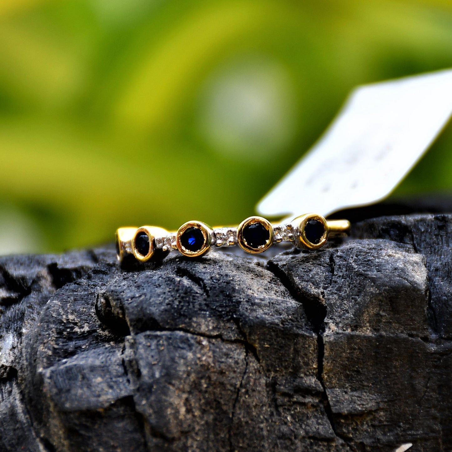 A 14kt gold diamond band ring with blue color stones, positioned on a rocky surface with green foliage in the background.