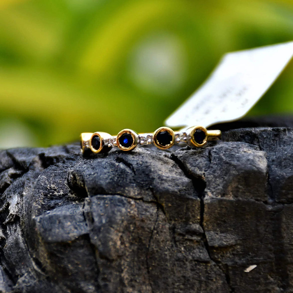 A 14kt gold diamond band ring with blue color stones, positioned on a rocky surface with green foliage in the background.