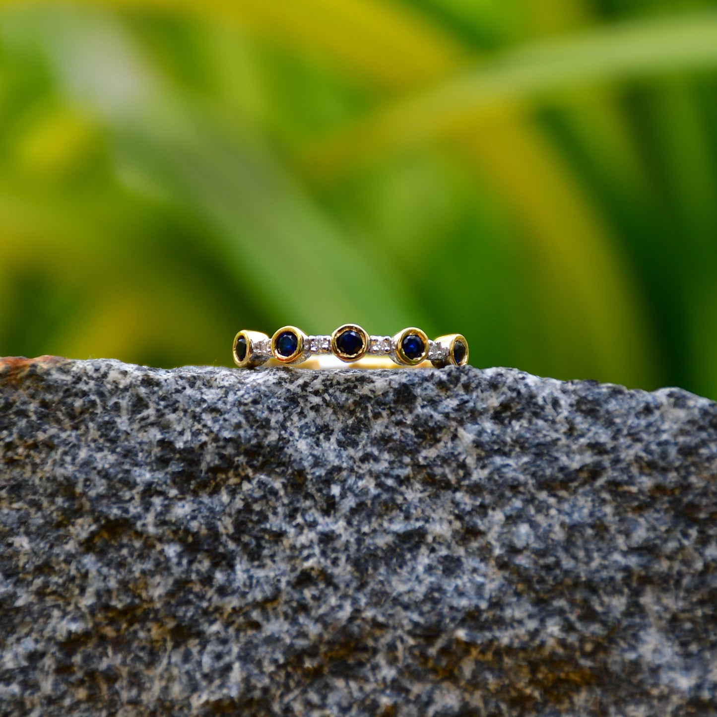 A 14kt gold diamond band ring with blue color stones, positioned on a rocky surface with green foliage in the background.