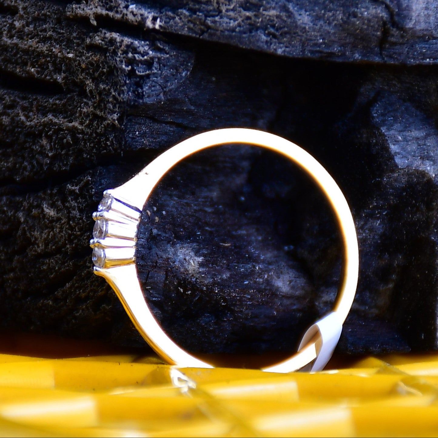 A 14kt gold diamond band ring with three brilliant cut natural diamonds on a yellow textured surface and against a rough black surface.