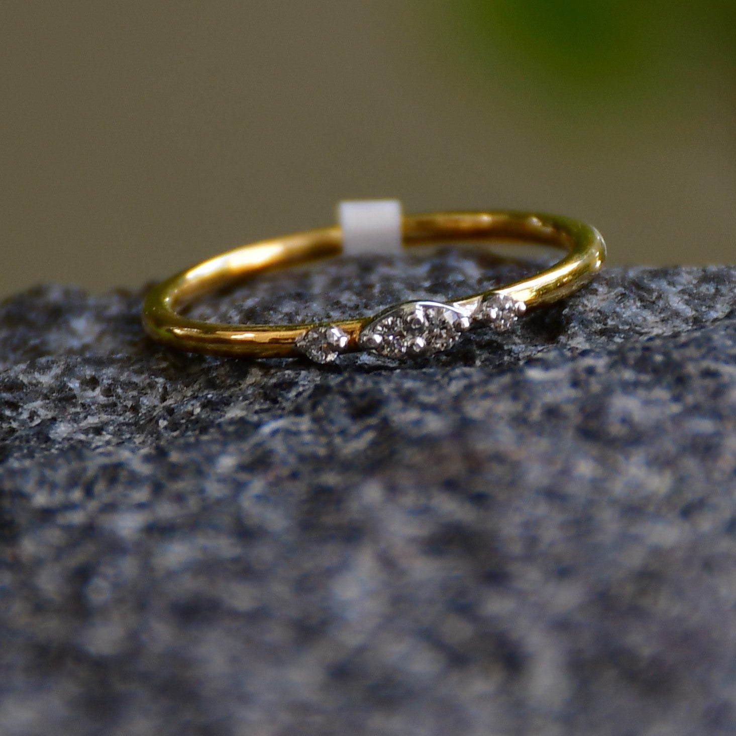 A close-up image of a delicate yellow gold ring featuring a small marquise-shaped cluster of diamonds at the center, displayed on a textured stone surface with a softly blurred natural background
