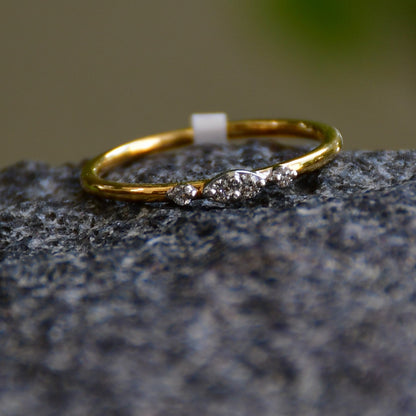A close-up image of a delicate yellow gold ring featuring a small marquise-shaped cluster of diamonds at the center, displayed on a textured stone surface with a softly blurred natural background