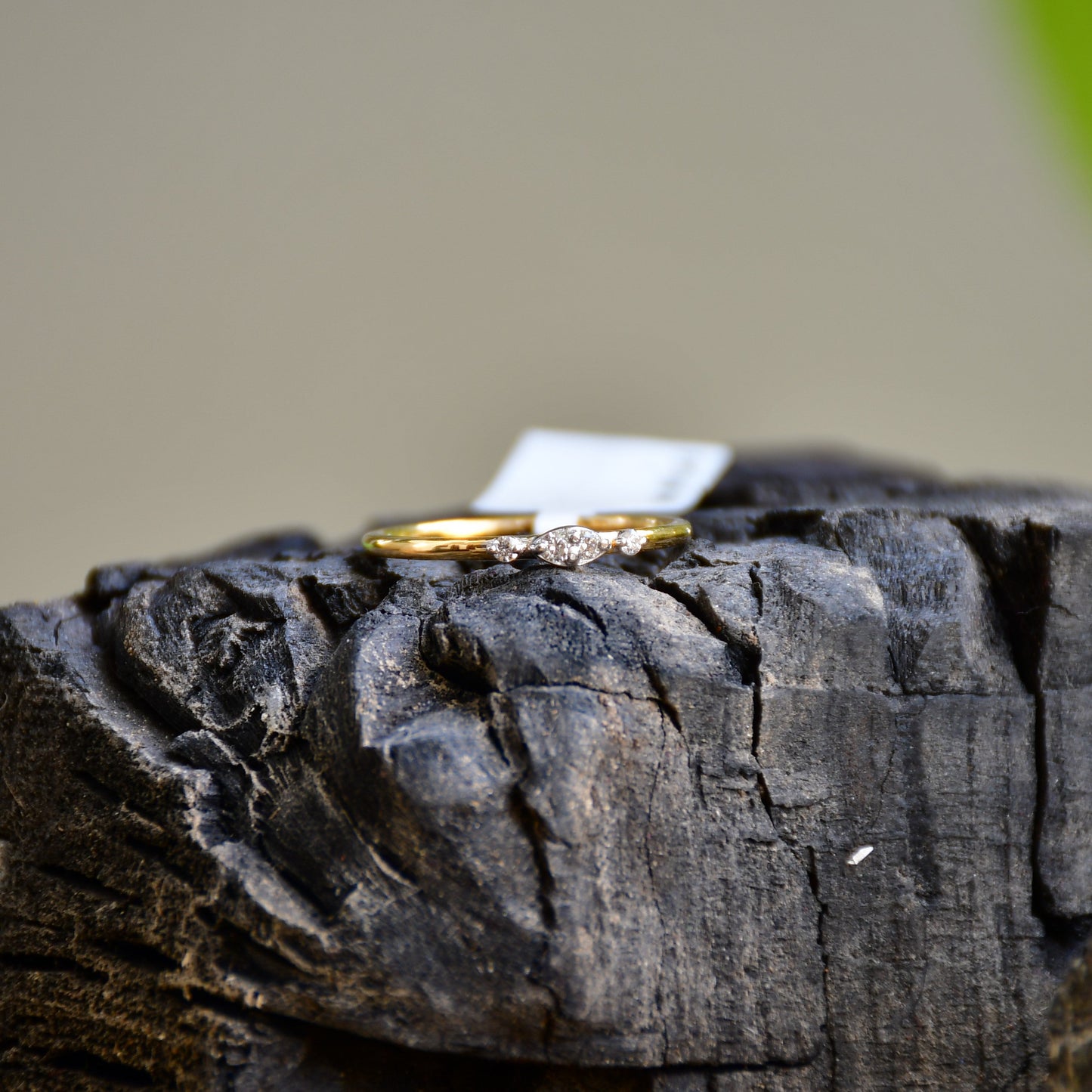 A close-up image of a delicate yellow gold ring featuring a small marquise-shaped cluster of diamonds at the center, displayed on a textured stone surface with a softly blurred natural background.