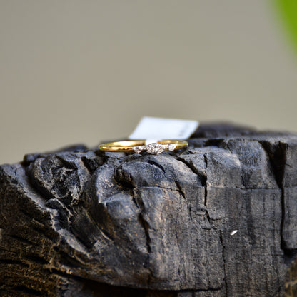 A close-up image of a delicate yellow gold ring featuring a small marquise-shaped cluster of diamonds at the center, displayed on a textured stone surface with a softly blurred natural background.