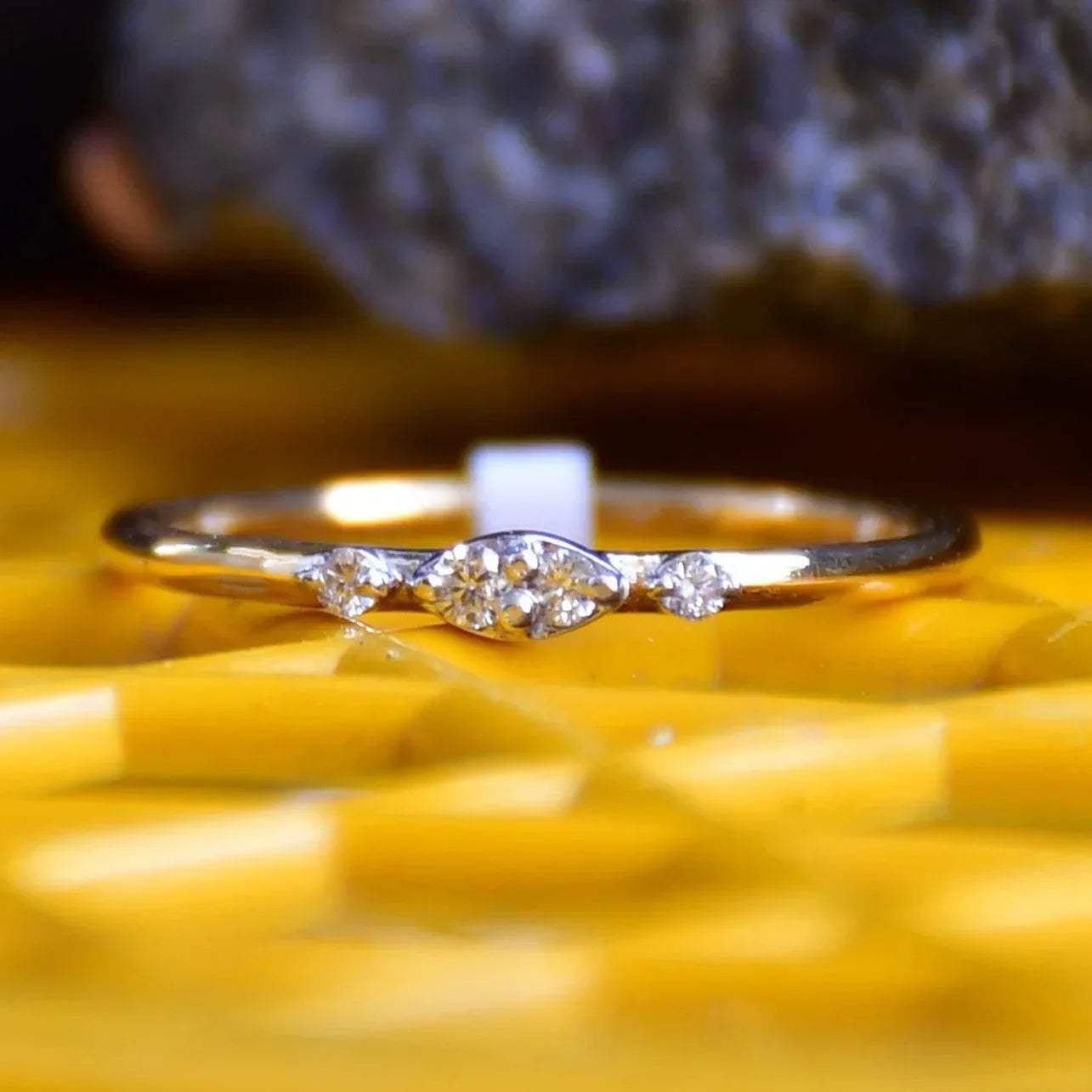 A close-up image of a delicate yellow gold ring featuring a small marquise-shaped cluster of diamonds at the center, displayed on a yellow surface with a softly blurred natural background