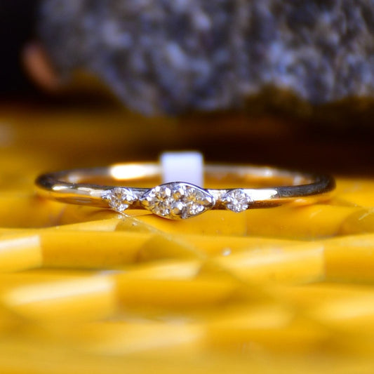 A close-up image of a delicate yellow gold ring featuring a small marquise-shaped cluster of diamonds at the center, displayed on a yellow surface with a softly blurred natural background
