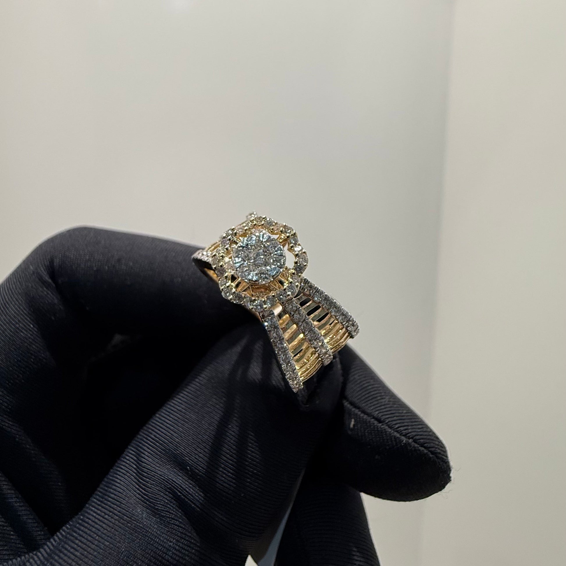 Elegant gold diamond ring featuring a round-cut central diamond surrounded by a halo of smaller diamonds, set on a split shank band adorned with rows of sparkling pavé diamonds, displayed on a black gloved hand against a neutral background.