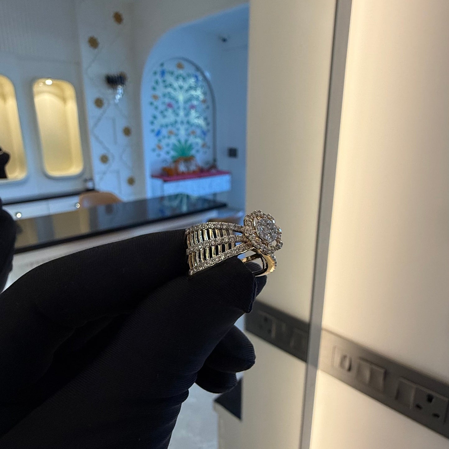 Elegant gold diamond ring featuring a round-cut central diamond surrounded by a halo of smaller diamonds, set on a split shank band adorned with rows of sparkling pavé diamonds, displayed on a black gloved hand against a neutral background.