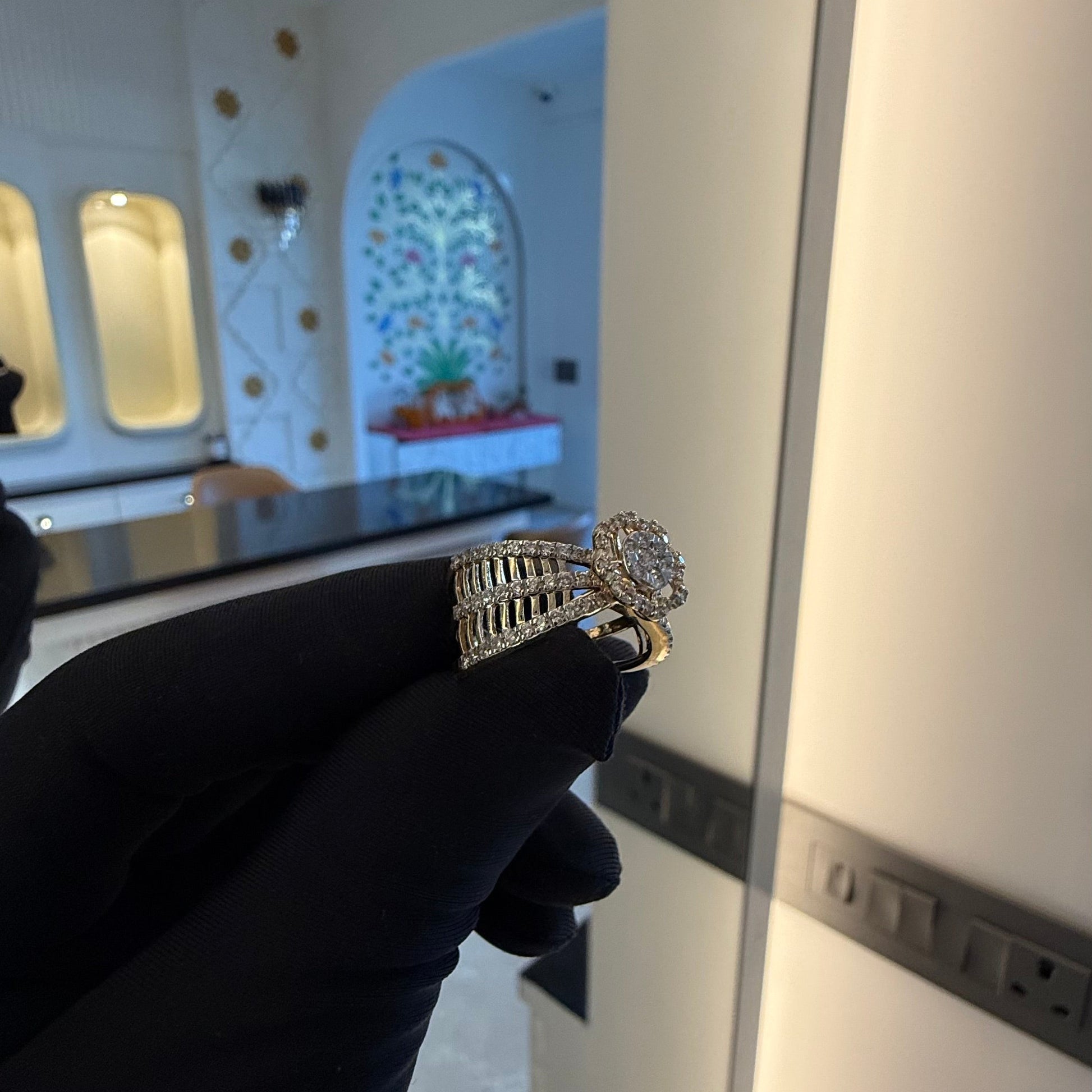 Elegant gold diamond ring featuring a round-cut central diamond surrounded by a halo of smaller diamonds, set on a split shank band adorned with rows of sparkling pavé diamonds, displayed on a black gloved hand against a neutral background.